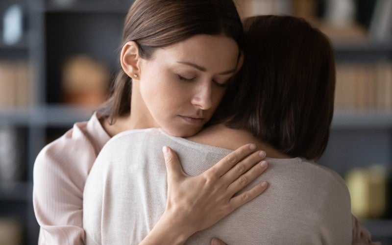 A young woman with closed eyes and a pained expression hugs another person with short dark hair, comforting them.