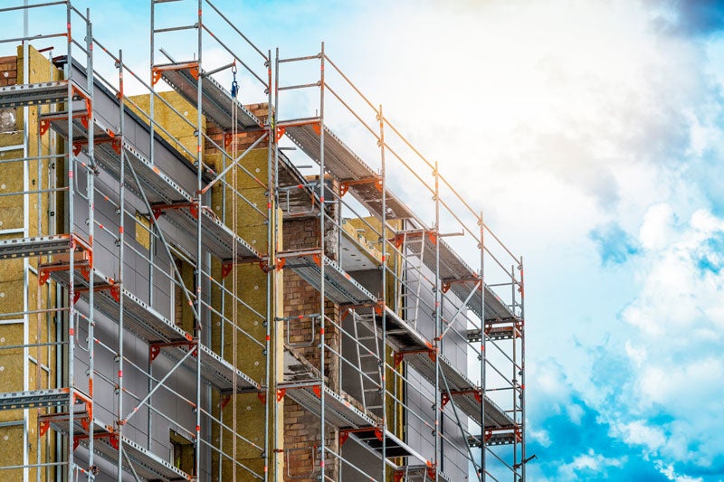 Scaffolding surrounds a building under construction, revealing insulation and brickwork on a sunny, cloudy day.