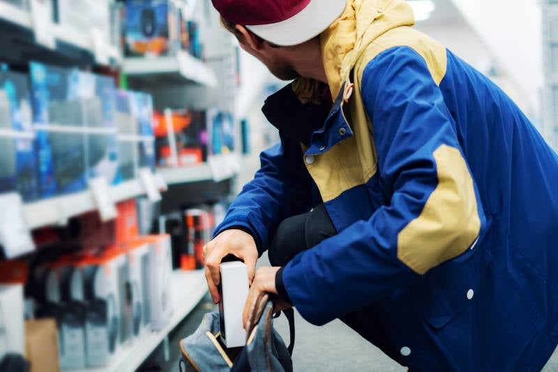 A person in a blue and yellow jacket and baseball cap stealthily placing a white box into a bag in a store aisle.