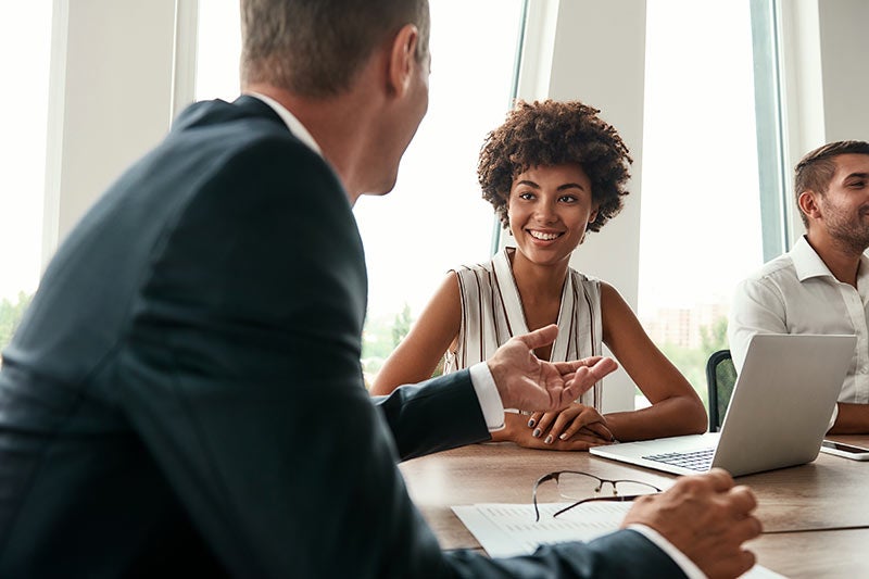 A Black woman with curly hair smiles at a man in a business suit across a conference table.