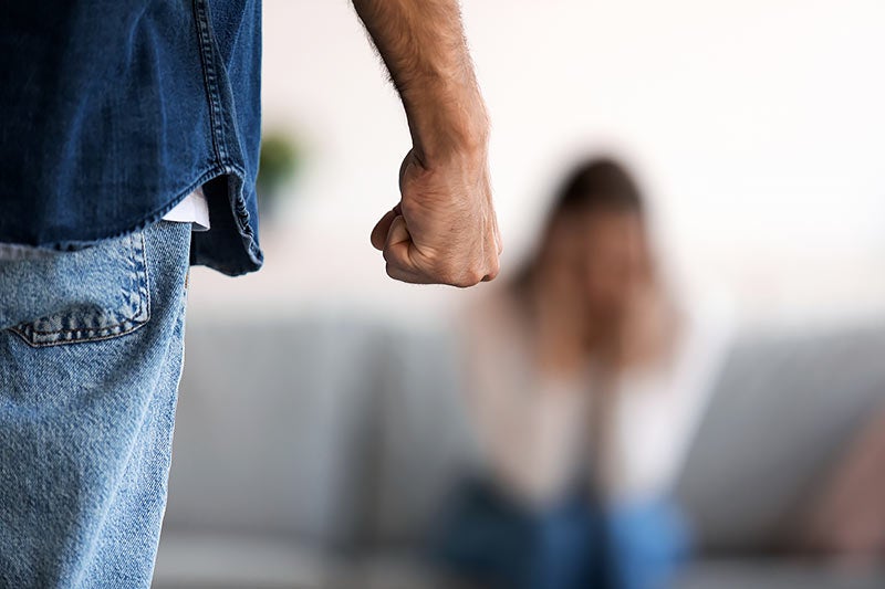 A person in jeans clenches their fist, standing in front of a blurred woman sitting on a sofa.