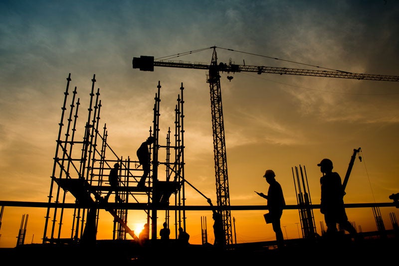 Silhouetted construction workers on scaffolding and a crane against a gradient orange and blue sky at sunset.