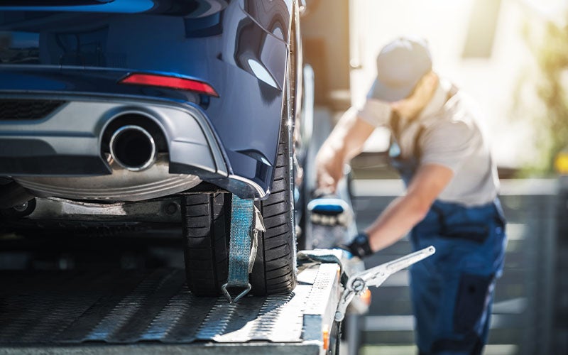 A blue car's rear wheel is strapped onto a tow truck by a blurred worker in work overalls, bright sun behind.