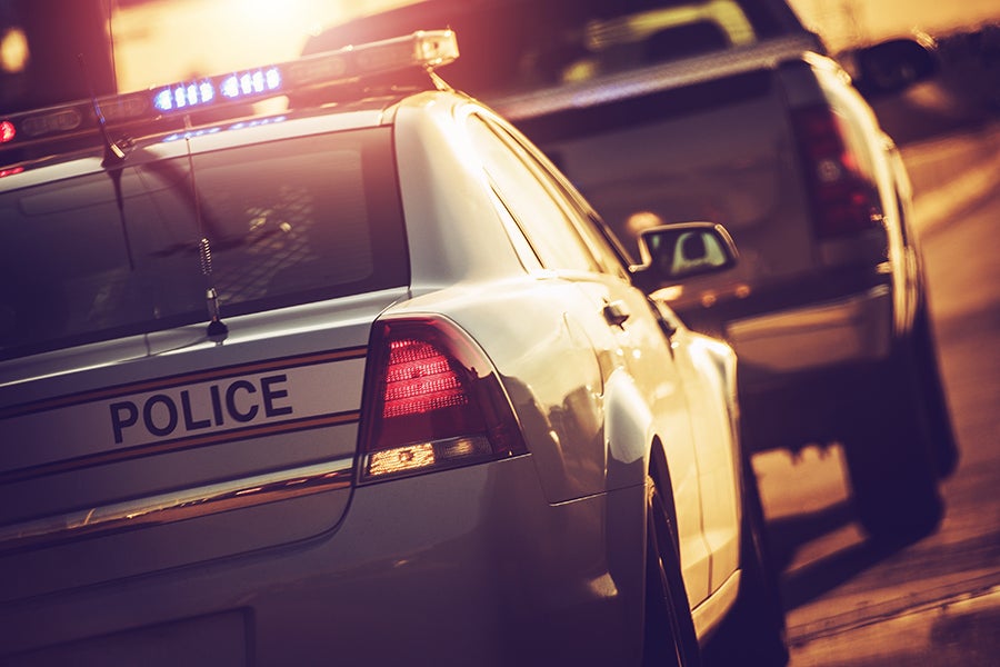 Rear view of a police car with flashing lights and a pursuit vehicle behind it under bright sunlight.