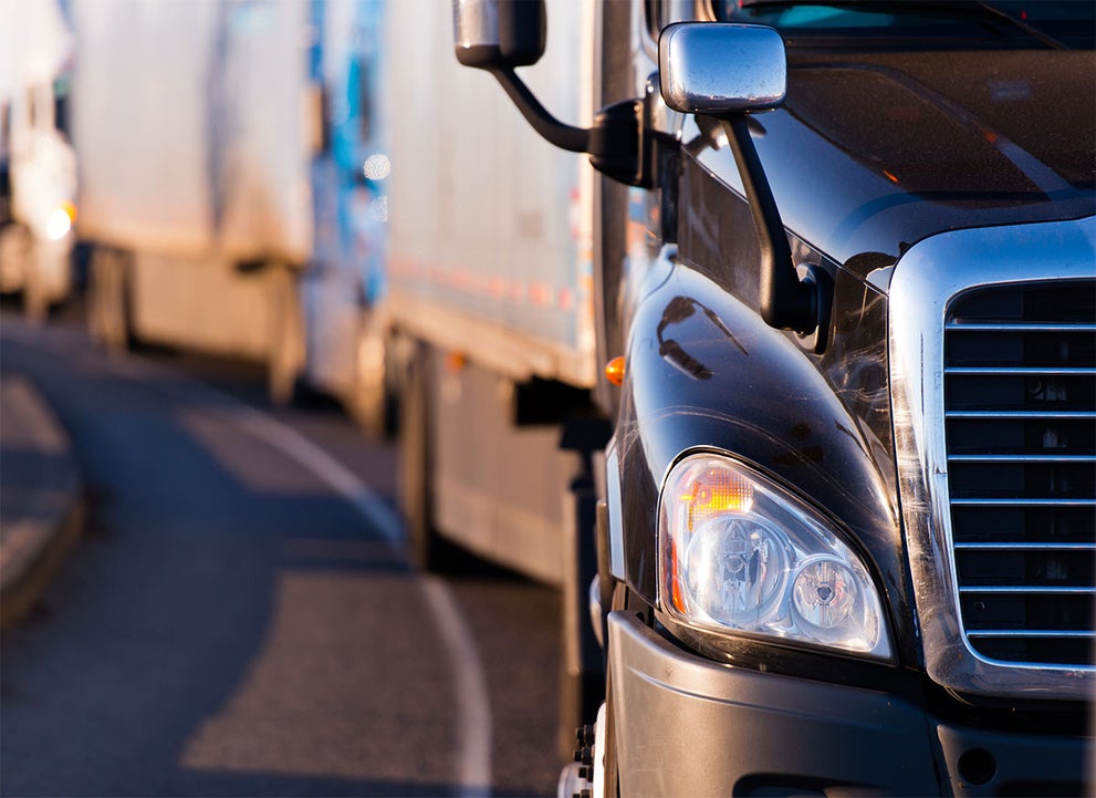 Close-up of a semi-truck's black hood, headlight, and chrome grille, with more trucks blurred in the background.