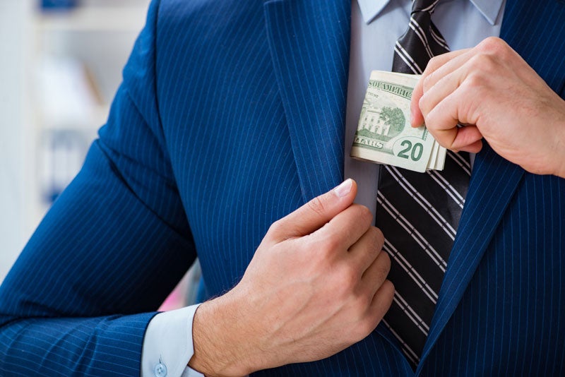 A person in a blue pinstripe suit and striped tie puts a wad of twenty-dollar bills into their inner jacket pocket.