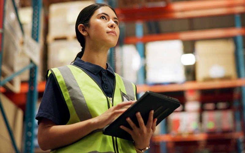 Woman in a high-visibility vest and dark polo shirt, looking up while holding a tablet in a warehouse.
