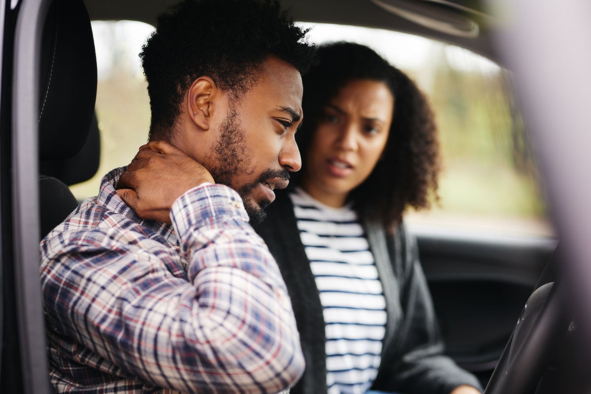 A man with a neck brace looks concerned while sitting next to a worried woman in a car.