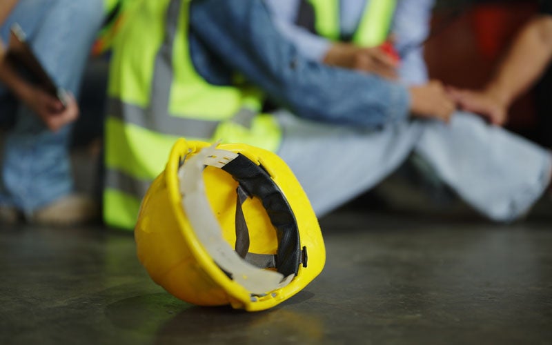 A yellow hard hat rests on the ground while two people in safety vests and jeans sit behind it.