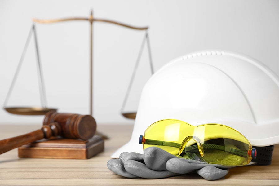 Gavel and scale of justice with a white hard hat, yellow safety glasses, and grey work gloves on a wooden desk.