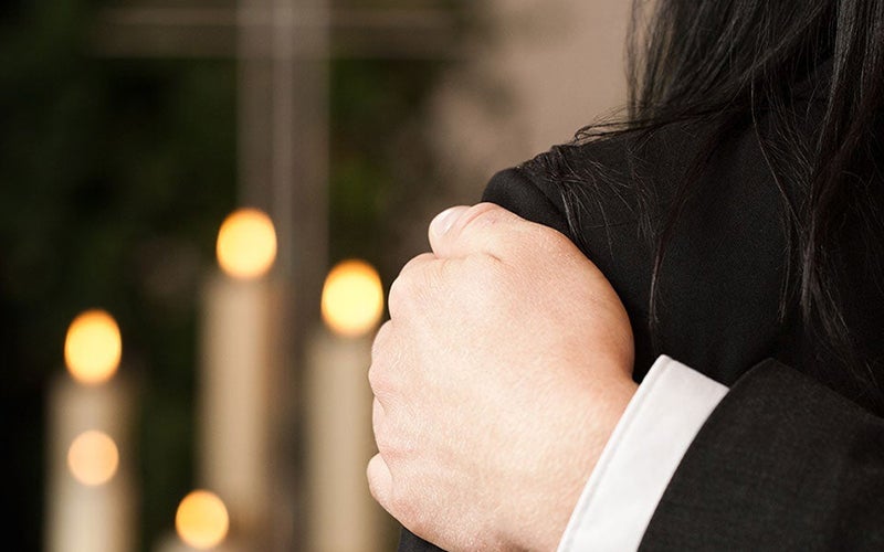 A hand comforting a person’s shoulder, with blurred lit candles in the background.
